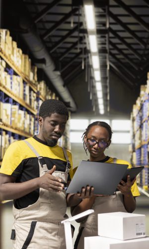 Black employees checking inventory and shipment details on laptop before applying awb tags. Large scale distribution operations with structured workflow and efficient merchandise tracking.